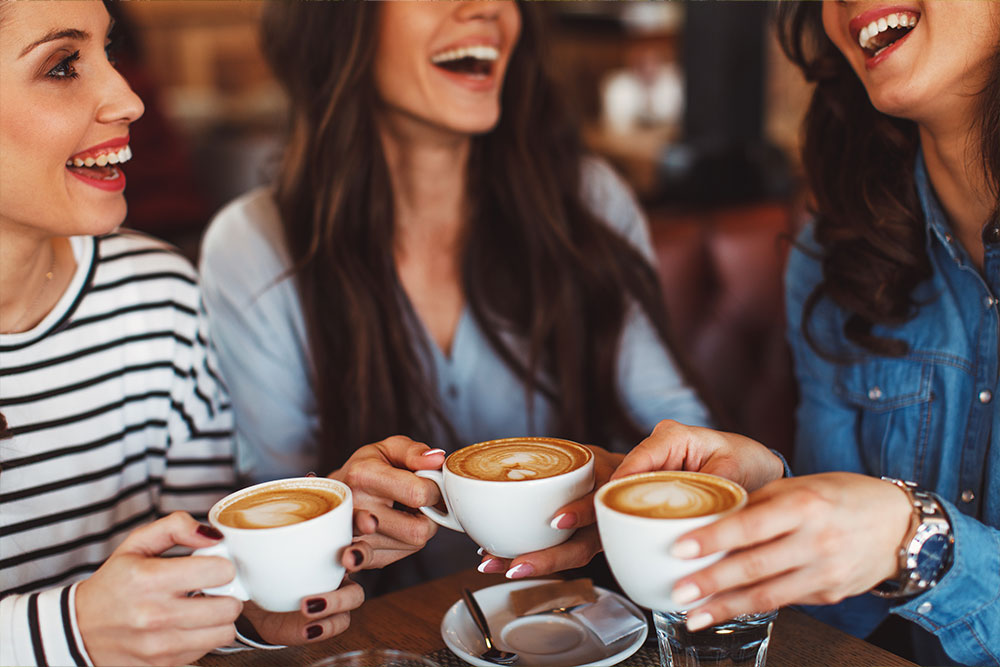 3 Freundinnen mit Kaffee in der Hand am Tisch in einem Café lachen herzlich.